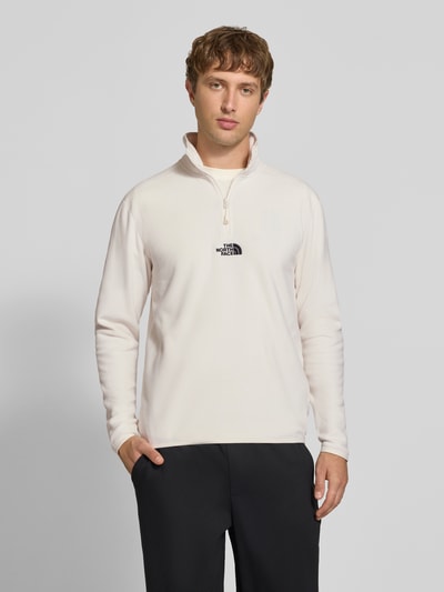 Männermodel trägt Fleecepullover mit Label-Stitching von The North Face in Creme, Stehkragen, Reißverschluss, Regular Fit, Ganzkörperansicht von vorne.