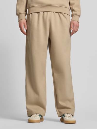 Mężczyzna nosi Wide Fit Sweatpants mit elastischem Bund od adidas Originals, bawełniany miks, beżowy kolor, logo, szeroki krój, widok z przodu, zdjęcie całej sylwetki.