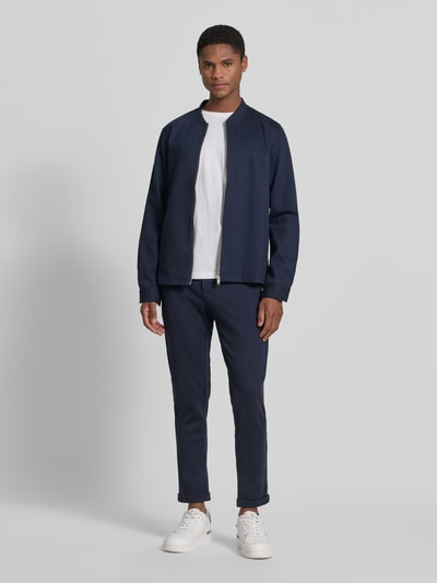 Męski model nosi Regular Fit Sweatjacke mit Stehkragen od Lindbergh z elastycznej mieszanki materiałów, zamek błyskawiczny, wpuszczane kieszenie, długość tyłu w rozmiarze M 69 cm, długość rękawów w rozmiarze M 66 cm, granatowa, gładka faktura, krój regular fit, stójka, Ganzkörperansicht von vorne