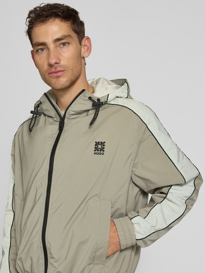 Männermodel trägt Regular Fit Jacke mit Kapuze Modell 'BAVO2621' von HUGO in Beige mit weißem Streifen, Logo-Detail, Nahaufnahme Oberkörper.