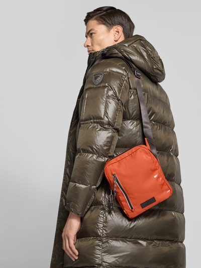 Männermodel trägt Crossbody Bag mit Statement-Print Modell 'EXPLORE' von camel active, rostorange, Außentasche mit Reißverschluss, Ganzkörperansicht Rückansicht.