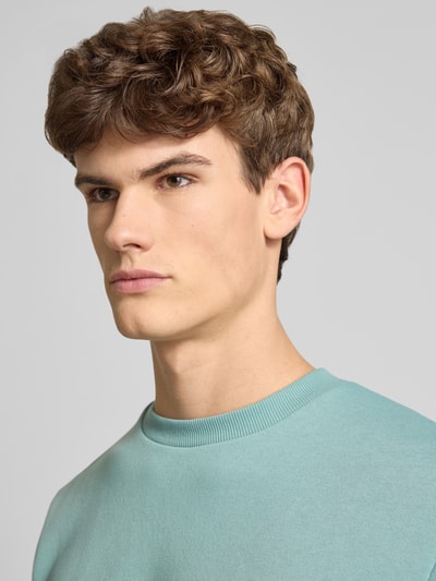 Męski model nosi Sweatshirt mit gerippten Abschlüssen Modell 'BRADLEY' od Jack & Jones, materiał z bawełną, krój regular fit, jasnoniebieski odcień, tekstura gładka, dekolt okrągły, zbliżenie na górną część ciała.