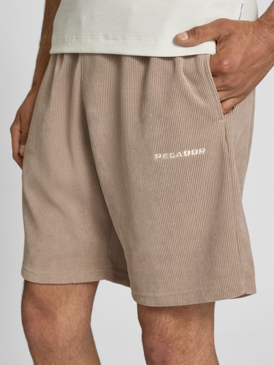 Männermodel trägt Relaxed Fit Shorts mit structuurpatroon model 'Lagos' van Pegador in beige elastische materiaalmix, zichtbare band, detailopname. Kontext: heren korte broek van Pegador, elastische materiaalmix, relaxed fit, model 'Lagos', structuurpatroon, elastische band, bandwijdte maat M: 38 cm, binnenbeenlengte maat M: 21 cm.
