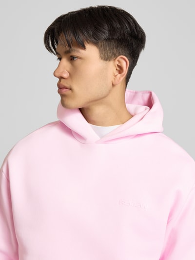 Männermodel trägt Hoodie mit Känguru-Tasche von REVIEW in Rosa, Baumwollmischung, Label-Detail, elastische Abschlüsse, Nahaufnahme Oberkörper.