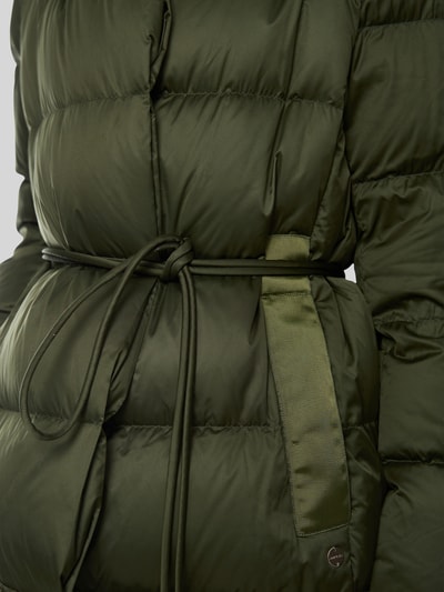 Frauenmodel trägt Steppmantel mit Bindegürtel von Marc Cain, olivgrün, glatte Steppstruktur, Eingrifftaschen, Nahaufnahme Oberkörper. Rückenlänge, Schulterbreite, Brustweite und Ärmellänge laut Größe 36 im Kontext.