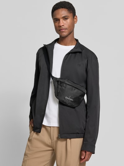 Männermodel trägt czarną saszetkę nerka z wyhaftowanym logo Bauchtasche mit Label Stitching Modell 'BOLD WAISTBAG' von CK Calvin Klein, Herren Umhängetasche; Ganzkörperansicht von vorne