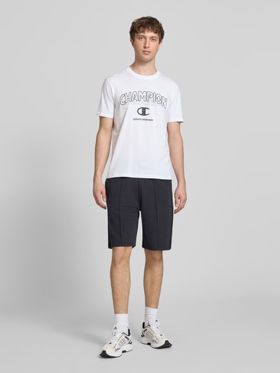 Männermodel trägt T-Shirt mit Label-Print und Rundhalsausschnitt von CHAMPION aus reiner Baumwolle, Regular Fit, Ganzkörperansicht von vorne.