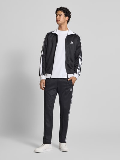 Männermodel trägt Trainingshose mit Label-Stitching Modell 'BECKENBAUER' von adidas Originals, Baumwoll-Mix, Regular-Fit, schwarze Farbe, elastischer Bund, Gesäßtasche, Innenbeinlänge 77 cm, Ganzkörperansicht von vorne.