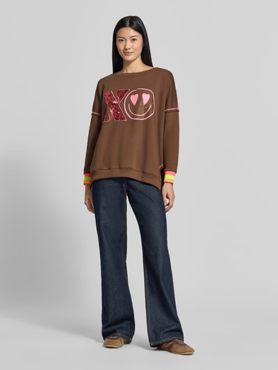 Frauenmodel trägt Oversized Sweatshirt aus reiner Baumwolle Modell 'XO Heartface' von miss goodlife, braun mit pinkem Statement-Print, Ziernähte, elastische Abschlüsse, Ganzkörperansicht von vorne.