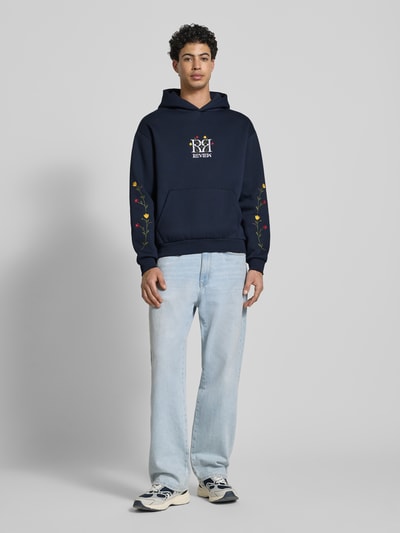 Männermodel trägt Regular Fit Hoodie mit Logo-Stitching von REVIEW, navy, florale Stickerei, Känguru-Tasche, Baumwoll-Mix, Ganzkörperansicht von vorne.