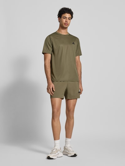 Männermodel trägt T-Shirt mit Label-Print von adidas Training in Oliv, Lockerer-Schnitt, gerippter Rundhalsausschnitt, Ganzkörperansicht von vorne. Rückenlänge bei Größe M: 70 cm.