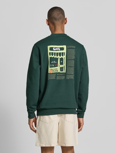 Männermodel trägt Sweatshirt mit Label-Details und Rundhalsausschnitt von Les Deux im Baumwoll-Elasthan-Mix, Regular-Fit, Rückenlänge 66 cm, Ärmellänge 70 cm, dunkelgrün mit grafichem Print, Ganzkörperansicht Rückansicht.