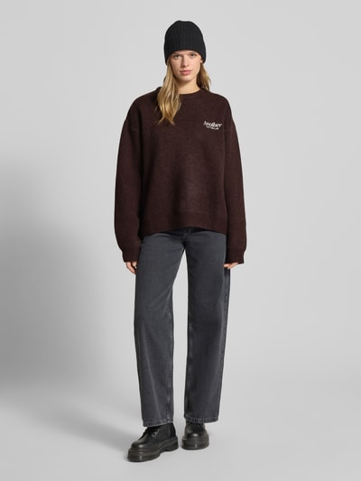 Frauenmodel trägt Oversized Strickpullover mit Rückenmotiv Modell 'Wine & Vibe' von ANOTHER COTTON LAB, braune Textur, Label-Stitching, Rundhalsausschnitt, Rückenlänge 62 cm, Ärmellänge 68 cm, Ganzkörperansicht von vorne.