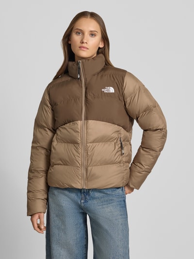 Frauenmodel trägt Steppjacke mit Reißverschlusstaschen Modell 'SAIKURU' von The North Face w kolorach brązu, krój regular fit, stójka, wyhaftowane logo, zamek błyskawiczny, kieszenie zapinane na zamek błyskawiczny, długość tyłu w rozmiarze S: 59 cm, szerokość ramion w rozmiarze S: 43 cm, szerokość w klatce piersiowej w rozmiarze S: 53 cm, długość rękawów w rozmiarze S: 62 cm, całe ciało, widok z przodu.