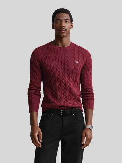 Männermodel trägt Regular Fit Pullover aus reiner Baumwolle von GANT in Dunkelrot mit Zopfmuster, Nahaufnahme Oberkörper. Herren Pullover.