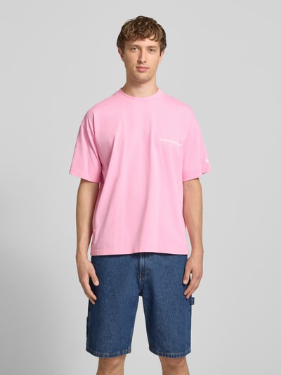 Męski model nosi T-Shirt mit Print und Rundhalsausschnitt von ANOTHER COTTON LAB, bawełna, krój relaxed fit, nadruk, okrągły dekolt, długość tyłu w rozmiarze M 69 cm, jasnoróżowy, gładka tkanina, niebieskie szorty, całopostaciowy widok z przodu.