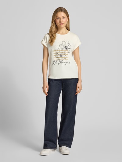 Modelka kobieca ma na sobie T-Shirt mit fixiertem Ärmelaufschlag Modell 'RINA' od Soyaconcept, bawełniany miks z elastanem, prosty krój, okrągły dekolt, nadruk z motywem, kremowy kolor, długość tyłu w rozmiarze S: 63 cm, widok całej sylwetki z przodu.