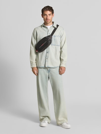 Męski model nosi czarno-czerwoną saszetkę nerkę Bauchtasche mit Logo-Detail Modell 'Ecodiver' von Samsonite; Główna przegroda z zamkiem błyskawicznym, regulowany pasek ze sprzączką, detal z logo; Szerokość w rozmiarze 1: 40 cm, Długość w rozmiarze 1: 17 cm, Głębokość w rozmiarze 1: 6 cm, Długość paska w rozmiarze 1: 94 cm; Ganzkörperansicht von vorne