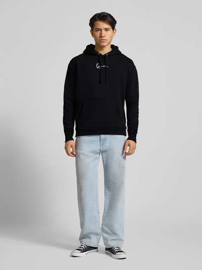 Männermodel trägt Hoodie mit Kapuze von KARL KANI in Schwarz aus Baumwoll-Mix, Label-Stitching, Känguru-Tasche, lockerer Schnitt, Ganzkörperansicht von vorne.