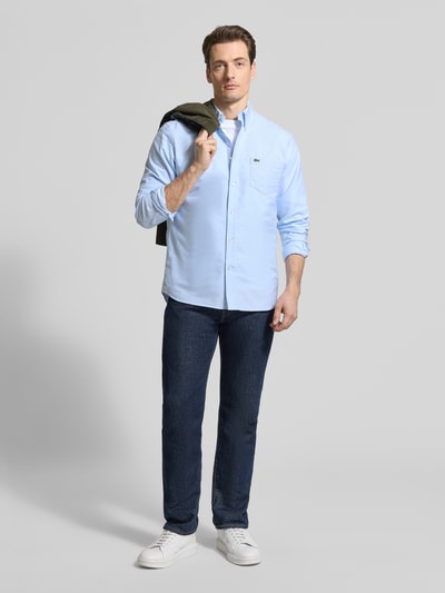 Męski model nosi Regular Fit Freizeithemd aus reiner Baumwolle od Lacoste w jednolitym jasnoniebieskim kolorze, z logo na kieszeni, długie rękawy, materiał bawełniany Oxford, widok na całą sylwetkę z przodu.