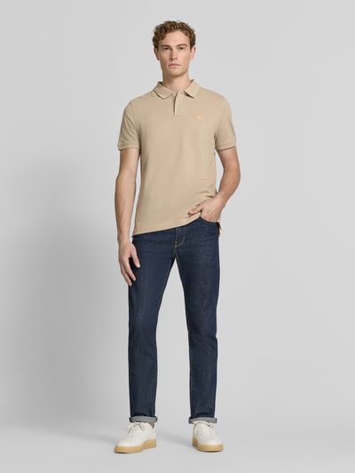 Męski model nosi Slim Fit Poloshirt mit Label-Stitching od MCNEAL z bawełny, krój slim fit, wyhaftowane logo, listwa guzikowa, długość tyłu 72 cm w rozmiarze M, kolor beżowy, ciemnoniebieskie spodnie, jasne buty, gładka tekstura, całopostaciowy widok z przodu.