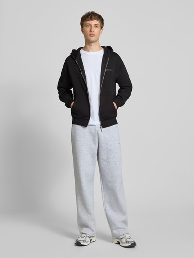 Männermodel trägt Relaxed Fit Sweatjacke aus Baumwoll-Mix mit Label-Detail von REVIEW in Schwarz, kombiniert mit heller Hose, Ganzkörperansicht von vorne. Rückenlänge 69 cm, Ärmellänge 61 cm, Kapuze, Känguru-Tasche, elastische Abschlüsse.