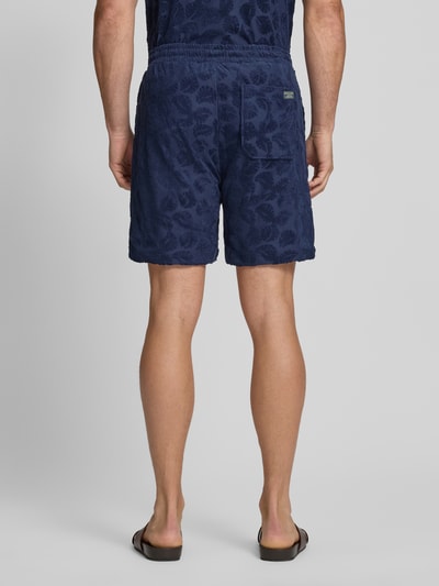 Męski model nosi Relaxed Fit Shorts aus Frottee mit Gesäßtasche od Scotch & Soda, mieszanka bawełny, krój relaxed fit, frota, elastyczny pas, wpuszczane kieszenie, kieszeń z tyłu, detal z logo, szerokość w rozmiarze M: 41 cm, długość wewnętrzna nogawki w rozmiarze M: 18 cm, granatowy, faktura froty, całkowity widok z tyłu.