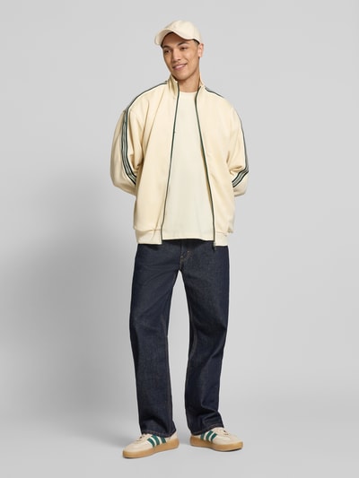 Männermodel trägt Relaxed Fit Sweatjacke mit Kontraststreifen Modell 'RIBER' von Only & Sons, Optik: Unifarben, Passform: Relaxed-Fit, Verschluss: Reißverschluss, Applikation: Kontraststreifen, Kragenform: Stehkragen, Ärmelform: Langarm, Ärmelabschluss: Gerippt, Rumpfabschluss: Gerippt, Taschen: Seitliche Eingrifftaschen, Materialverarbeitung: Sweat, helle Farbtöne, glatte Textur, Ganzkörperansicht von vorne.