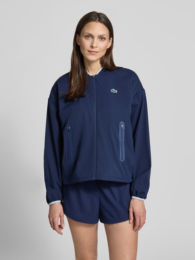 Frauenmodel trägt Regular Fit Trainingsjacke mit Baseball-Kragen von Lacoste Sport, unifarben dunkelblau, elastische Ärmelabschlüsse, Sicht auf Reißverschlusstaschen, Nahaufnahme Oberkörper von vorne