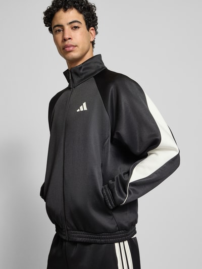 Męski model nosi Kurtka treningowa z logo i stójką od adidas Sportswear, mieszanka materiałów z dodatkiem bawełny, krój regular fit, logo, stójka, zamek błyskawiczny, długość tyłu 68 cm, długość rękawa 67 cm, kolor czarny z białymi akcentami, gładka tekstura. Ujęcie: widok całej sylwetki z przodu.