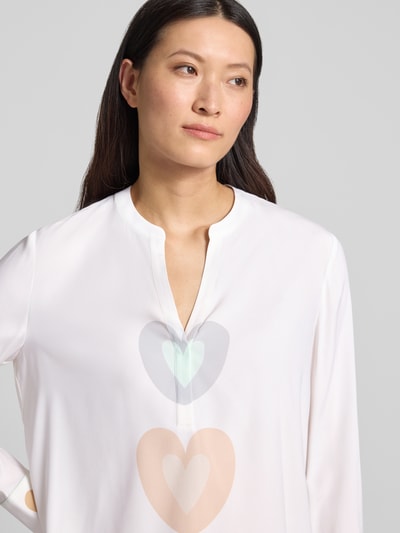 Modelka kobieca ma na sobie Relaxed Fit Tunika-Bluse mit Allover-Motiv-Print od HERZENS, biały kolor, dekolt z lamówką, nadruk z motywem na całej powierzchni, tunika z długim rękawem, przedłużony tył, długość tyłu w rozmiarze 36: 68 cm, długość rękawów w rozmiarze 36: 63 cm, widok z przodu, zbliżenie na górną część ciała.