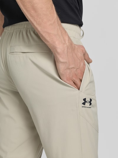 Männermodel trägt Regular Fit Jogger mit Logo-Print Modell 'Vibe' van Under Armour, beige, elastische taille en zakken, detailopname heup, heren jogger, elastische materialenmix, regular fit, logo-print, elastische tailleband met trekkoord, steekzakken, elastische pijpuiteinden, binnenbeenlengte maat M 78 cm.