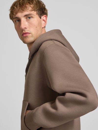 Männermodel trägt Relaxed Fit Sweatjacke mit Zweiwege-Reißverschluss von JACK & JONES in Beige, glatte Textur, Nahaufnahme Oberkörper. Elastischer Material-Mix, Känguru-Tasche und elastische Abschlüsse sichtbar.