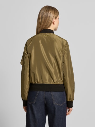 Frauenmodel trägt Bomberjacke mit Label-Patch und Reißverschlusstasche am Ärmel von Original Bombers in Olivgrün mit Stehkragen, Lockerer-Schnitt, Reißverschluss, Logo-Stitching, elastischen Abschlüssen, und seitlichen Eingrifftaschen mit Druckknopfverschluss, Rückenlänge 53 cm, Schulterbreite 37 cm, Brustweite 48 cm, Ärmellänge 63 cm, Ganzkörperansicht Rückansicht.