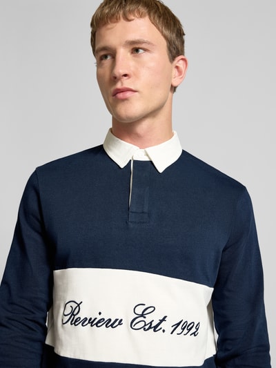 Männermodel trägt Regular Fit Langarm-Poloshirt mit Label-Stitching von REVIEW, Dunkelblau mit weißem Kontraststreifen, Reine Baumwolle, verdeckte Knopfleiste, Nahaufnahme Oberkörper. Rückenlänge 68 cm, Ärmellänge 72 cm, Größe M.