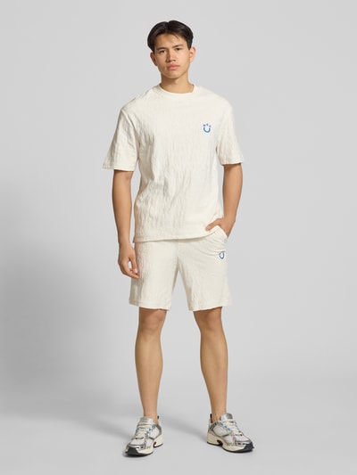 Männermodel trägt Regular Fit Shorts aus Frottee von HUGO in Creme mit Frottee-Optik, Tunnelzug, Logo-Stitching, seitliche Eingrifftaschen, aufgesetzte Tasche hinten, Ganzkörperansicht von vorne.