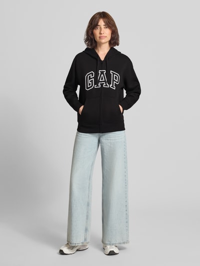 Vrouwenmodel draagt zwarte sweatjack met capuchon en kangoeroezak Model 'HERITAGE' van GAP, katoenmix, relaxed fit, logo, ritssluiting. Ganzlichaamsaanzicht van voren