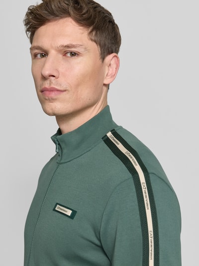 Männermodel trägt Sweatjacke mit Stehkragen und Zweiwege-Reißverschluss von EA7 Emporio Armani in Grün, Nahaufnahme Oberkörper. Regular Fit, Label-Detail, Kontraststreifen auf den Ärmeln.