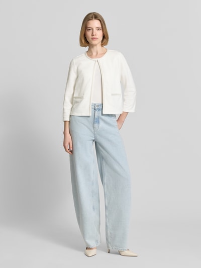Modelka kobieca nosi Regular Fit Jacke mit Zierbordüre od More & More, kremowa, tekstura elastyczna z bawełną, otwarty fason, przód z ozdobną lamówką, długość tyłu 48 cm, długość rękawa 47 cm, szerokie jasnoniebieskie spodnie, widok całej sylwetki z przodu.