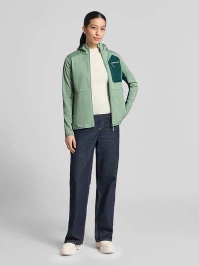 Frauenmodel trägt Jacke mit Logo-Stitching von PEAK PERFORMANCE aus Elastischer-Material-Mix mit körpernaher Passform, Kapuze, Reißverschluss und Brusttasche, Farbkontrast in Grün und Dunkelgrün, Ganzkörperansicht von vorne.