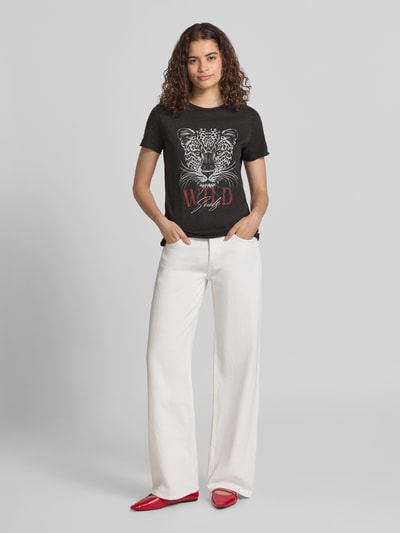 Frauenmodel trägt Regular Fit T-Shirt aus reiner Baumwolle 'ONLLUCY' von ONLY, schwarzes Shirt mit weißem Leopardenprint, weiße Hose, rote Schuhe, Ganzkörperansicht von vorne, Damen T-Shirt von ONLY.