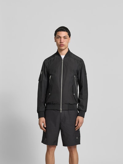 Männermodel trägt Bomberjacke mit Zweiwege-Reißverschluss von Moose Knuckles, schwarze Farbe, lockere Passform, Ganzkörperansicht von vorne. Rückenlänge 67 cm, Schulterbreite 46 cm, Ärmellänge 68 cm, Reißverschluss- und Ärmeltaschen, Stehkragen, elastische Abschlüsse.