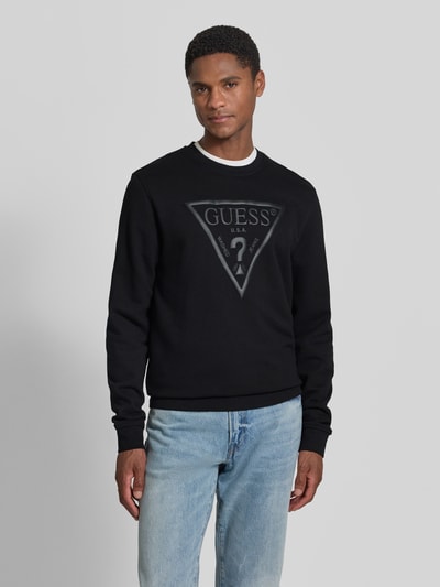 Männermodel trägt Regular Fit Sweatshirt mit Logo-Detail Modell 'Audley' von Guess aus Baumwoll-Mix in Schwarz, Ganzkörperansicht von vorne. Rückenlänge 69 cm, Ärmellänge 69 cm.