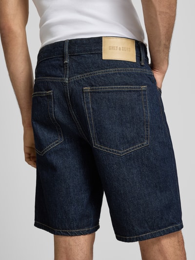 Männermodel trägt Regular Fit Jeansshorts aus reiner Baumwolle Modell 'EDGE' von Only & Sons in dunkler Denim-Optik mit Label-Patch, 5-Pocket-Design und Twill-Materialverarbeitung, Ganzkörperansicht Rückansicht.