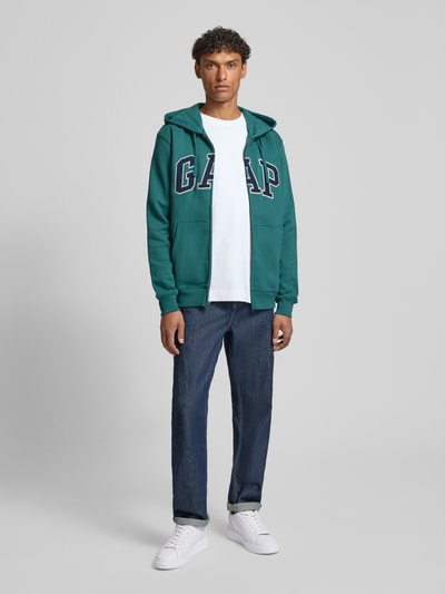 Männermodel trägt Sweatjacke mit Logo und Känguru-Tasche von GAP in Grün, Baumwoll-Mix, Regular Fit, Ganzkörperansicht von vorne.