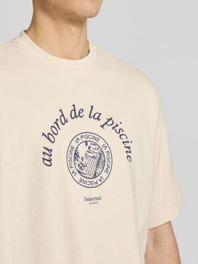 Männermodel trägt Relaxed Fit T-Shirt aus reiner Baumwolle Modell 'BOXYNORMAN GRAPHIC' von SELECTED HOMME in Beige mit blauem Motiv-Print, Jersey-Textur, Nahaufnahme Oberkörper.