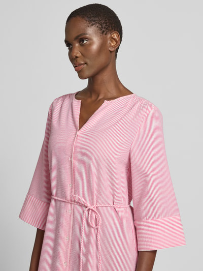 Modelka kobieca ma na sobie sukienkę koszulową z mieszanki bawełny, wiązanym paskiem i rękawami kimono B.Young Knielanges Hemdblusenkleid mit Bindegürtel Modell 'IME', długość do kolan, dekolt serafino, listwa guzikowa, zaokrąglony dół, kolor biało-różowy z cienkimi paskami, taliowany fason, szerokość w rozmiarze 36: 42 cm, długość tyłu: 103 cm, długość rękawów: 42 cm, ujęcie z przodu do pasa.