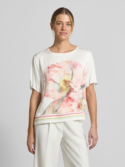 Modelka nosi Relaxed Fit T-Shirt aus reiner Viskose mit floralem Print od Betty Barclay, biała i różowa kolorystyka, wzór kwiatowy, okrągły dekolt, elastyczny dół, długość tyłu 57 cm, długość rękawa 24 cm przy rozmiarze 36, widok całej sylwetki z przodu.