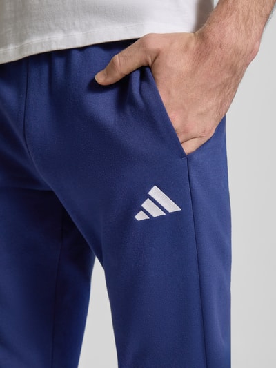 Männermodel trägt Trainingsanzug mit Label-Details von adidas Sportswear in Blau mit Eingrifftaschen, Detailaufnahme Oberschenkel.