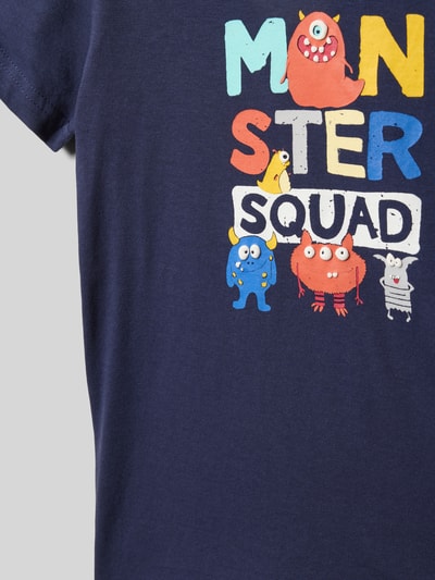 Mädchen trägt dziecięcy t-shirt Regular Fit T-Shirt mit Motiv-Print von Blue Seven aus bawełna, nadruk z motywem, okrągły dekolt, długość tyłu w rozmiarze 116: 44 cm, z kolorowym nadrukiem na ciemnoniebieskim tle, nahaufnahme górnej części torsu.